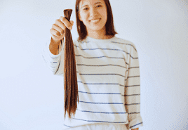 A woman smiling, holding a long cut ponytail against a plain background.